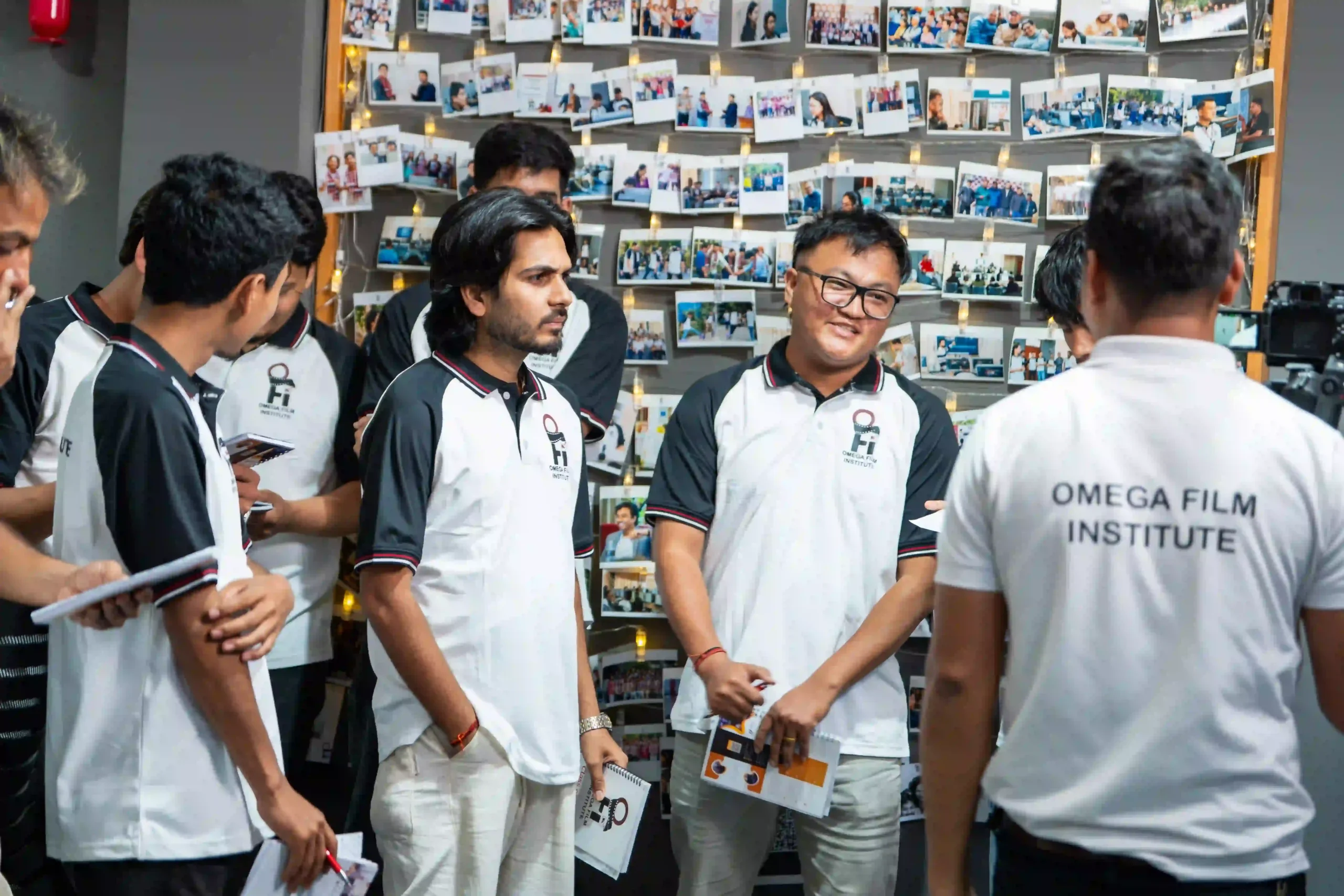 Group of young men in matching white and black Omega Film Institute shirts talking in front of a wall covered with photos and string lights.
