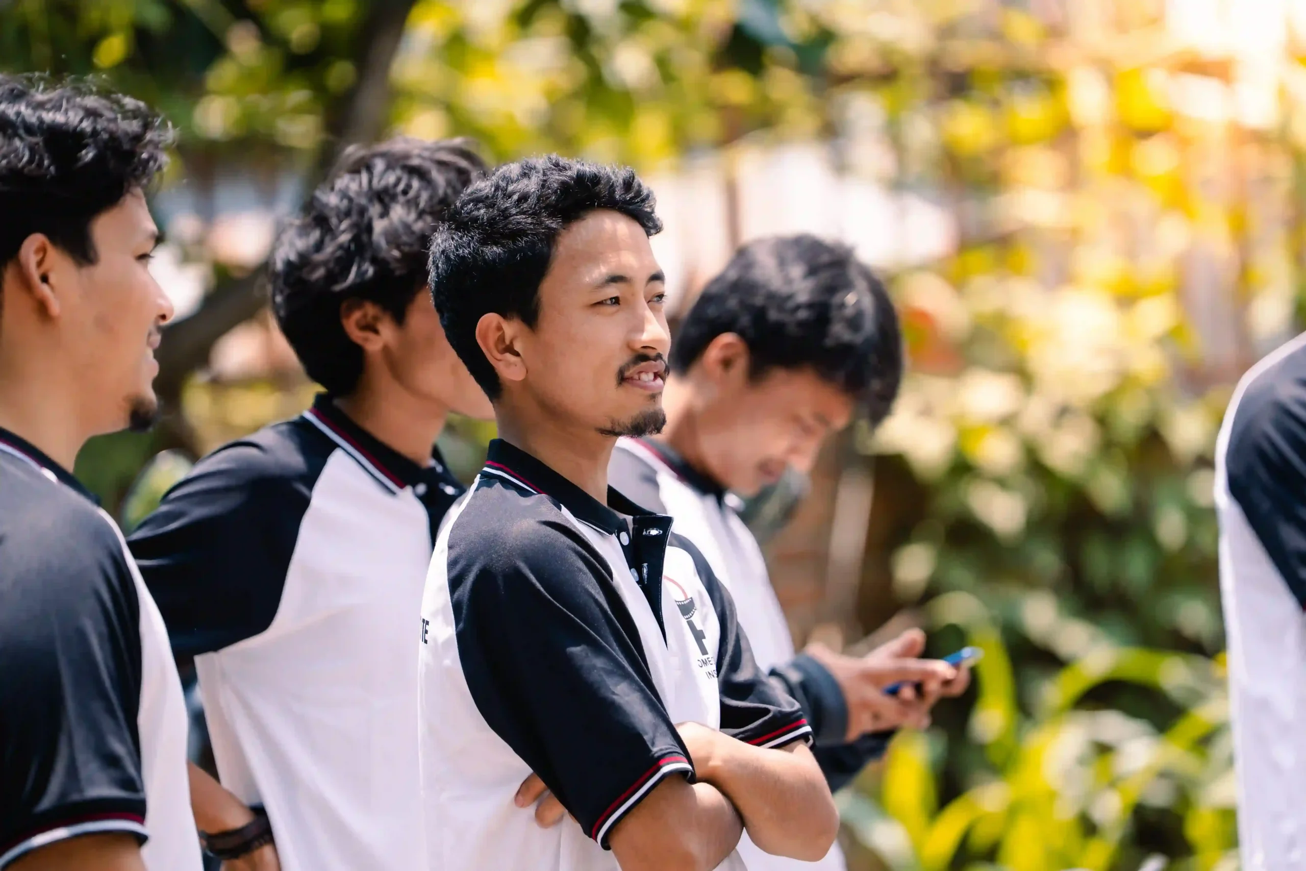 Group of men in matching black-and-white uniforms standing outdoors, arms crossed and smiling in a sunny park.