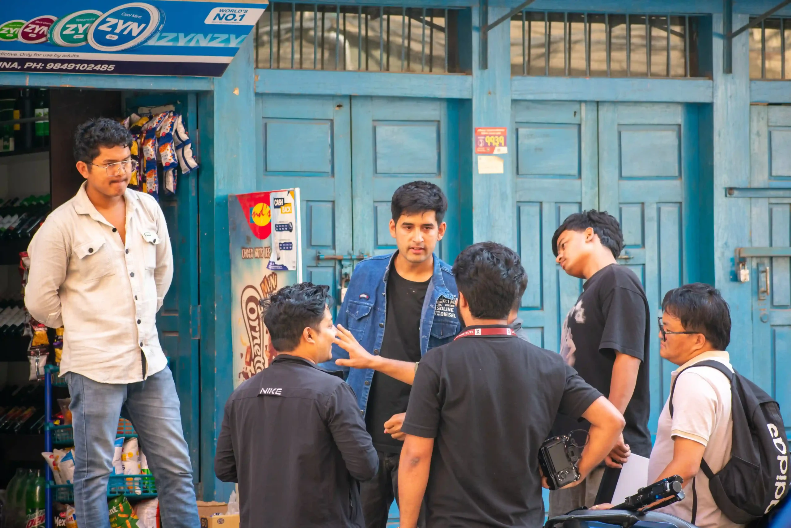 Group of young men stand and chat outside a blue storefront, one holding a camera bag nearby.