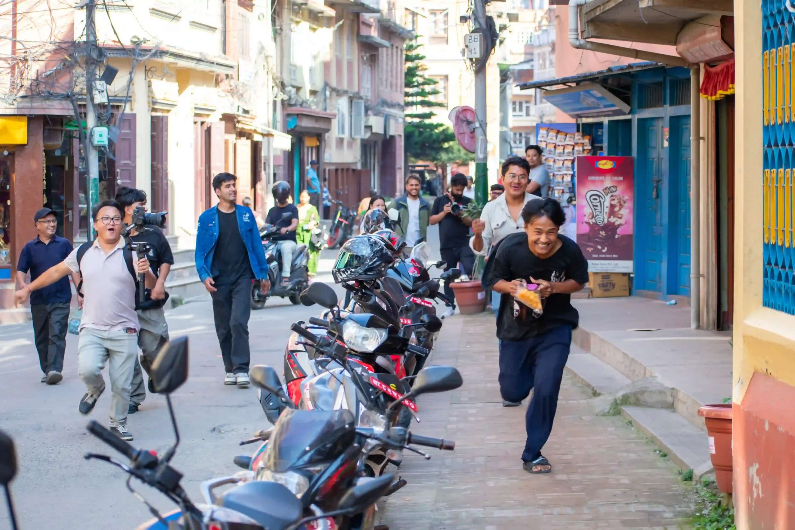 Group of people filming a street scene in a crowded alley; motorcycles parked along both sides.