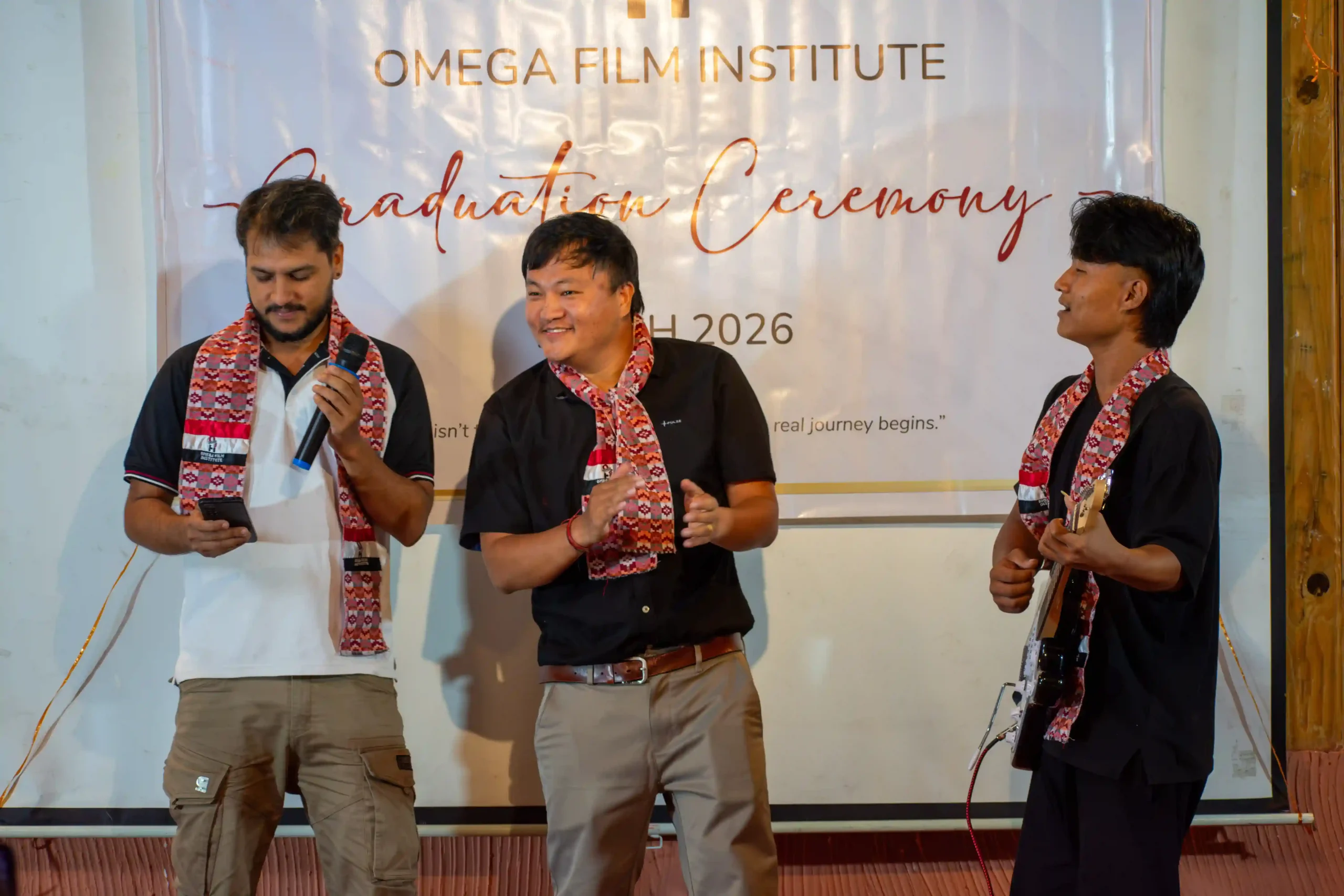 Three men in black shirts with patterned scarves stand on a stage during a graduation ceremony under an Omega Film Institute banner. One holds a microphone and phone, another smiles, the third holds a guitar/strap.