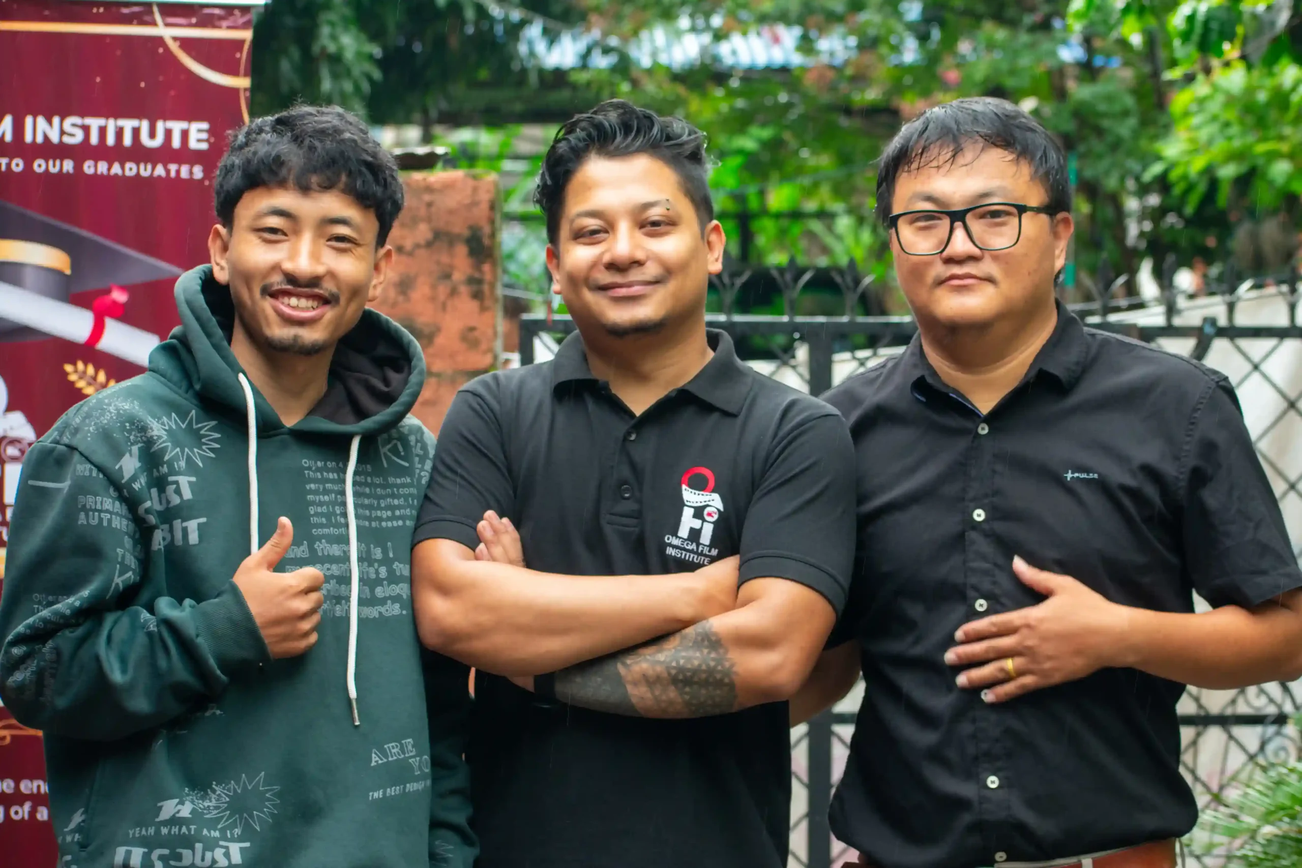 Three men pose together outdoors, smiling with arms crossed; left in a teal hoodie, middle in a black polo, right in a black shirt with glasses, greenery and a fence in the background.